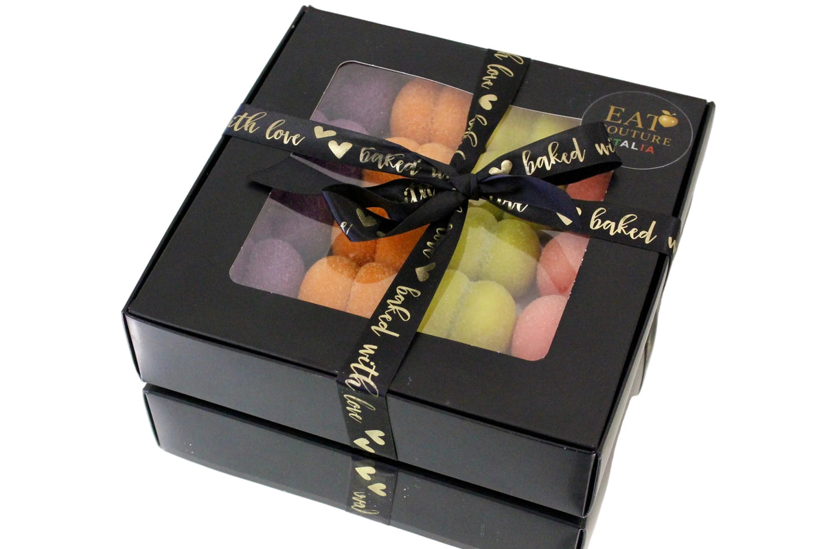 A square black gift box with two clear windows showing colorful macarons inside, tied with a black ribbon printed with gold text and hearts. A round sticker on the lid reads 'EAT GULF FOODS LLC'.