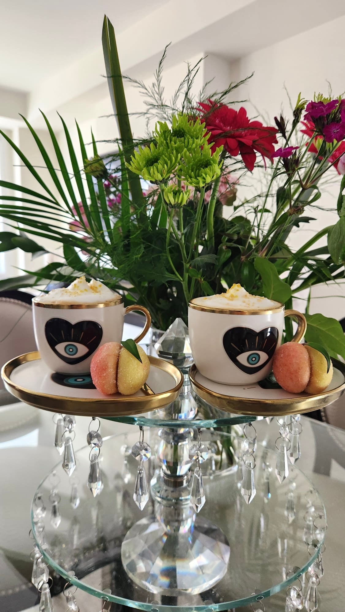 Two cups with heart designs on a glass stand with flowers and pastries in the background.