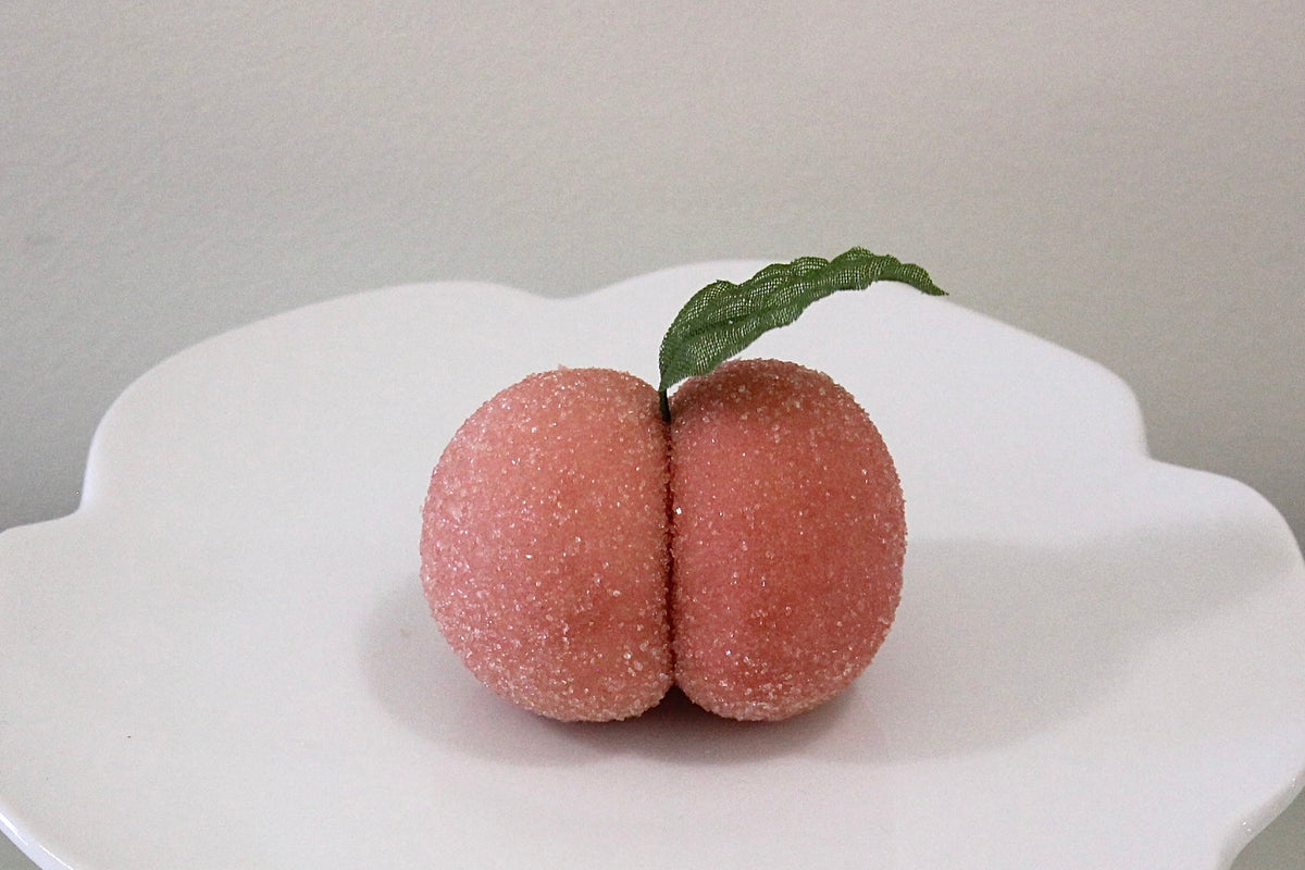 Two pinkish-brown, textured novelty items shaped like a pair of buttocks sit on a white pedestal plate with a small green leaf on top, set against a neutral gray background.