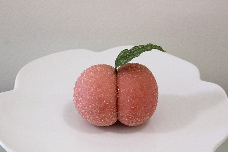Two pinkish-brown, textured novelty items shaped like a pair of buttocks sit on a white pedestal plate with a small green leaf on top, set against a neutral gray background.