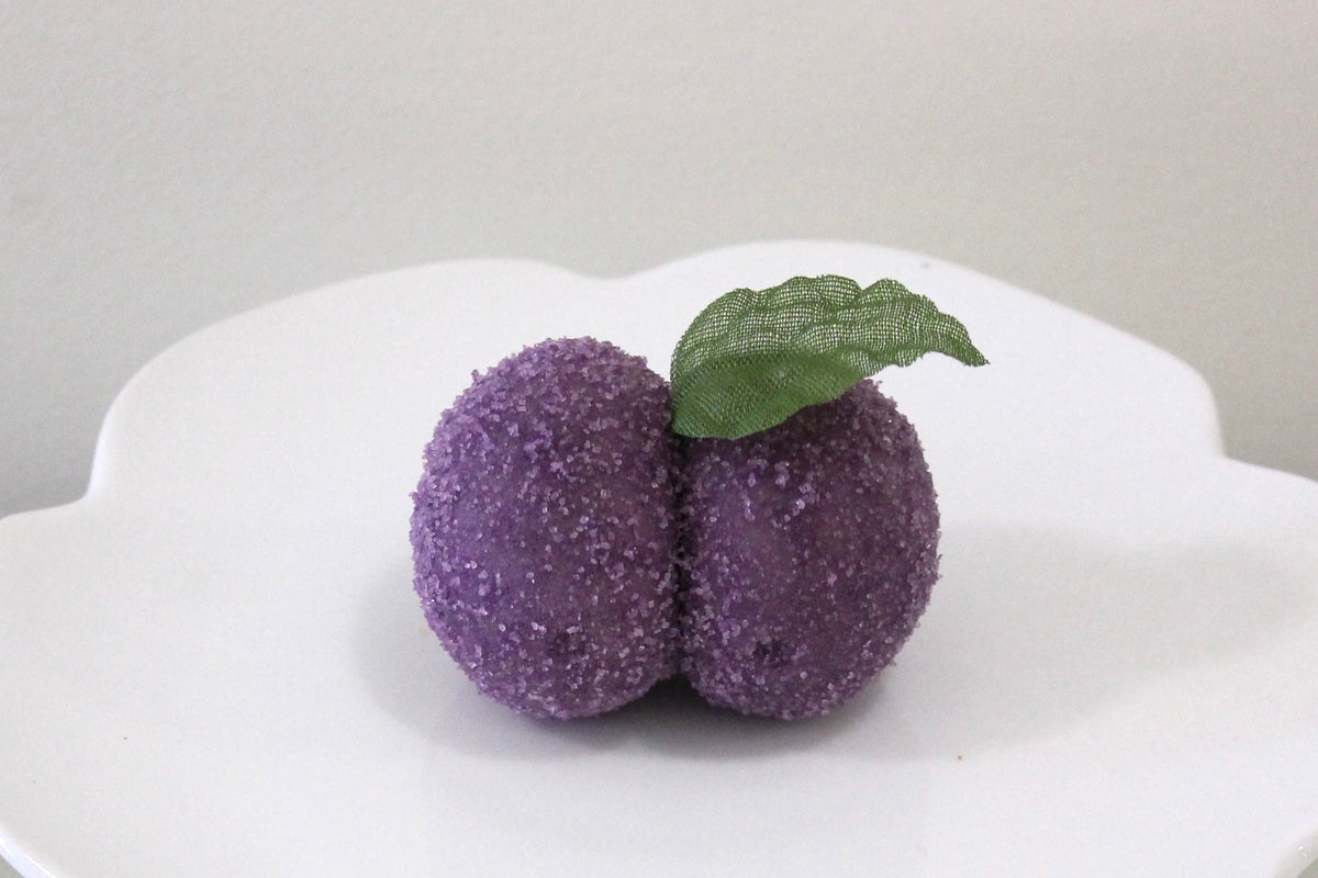 Two small purple pom-pom fruit decorations with a green leaf on top, displayed on a white ceramic pedestal plate against a neutral light gray background.