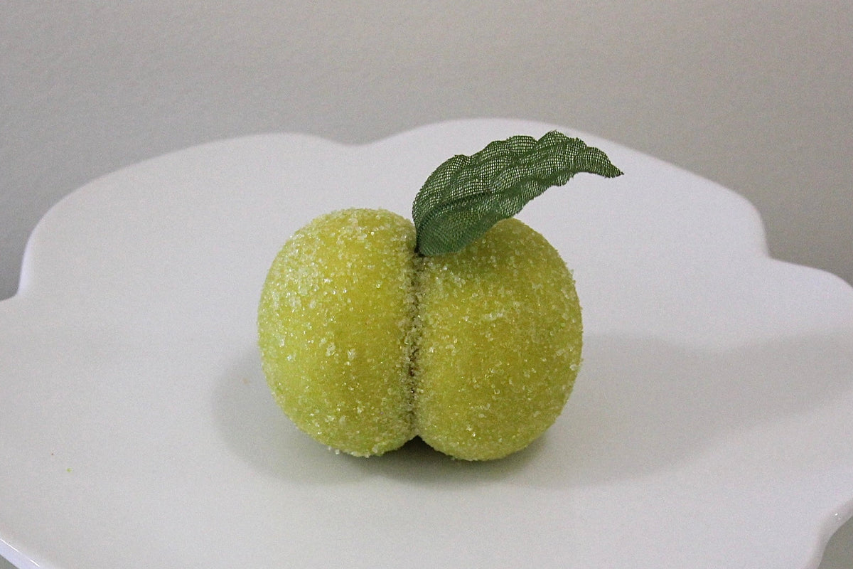 A white ceramic pedestal plate with a scalloped rim, holding two small green fruit-like decorations with a leaf on top, set against a neutral gray background.