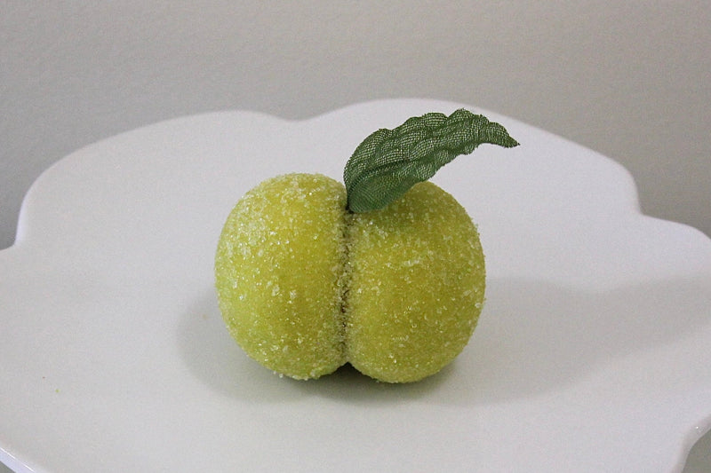 A white ceramic pedestal plate with a scalloped rim, holding two small green fruit-like decorations with a leaf on top, set against a neutral gray background.