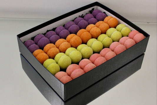 A black gift box filled with rows of colorful macarons in purple, orange, yellow, and pink, photographed on a light gray surface.