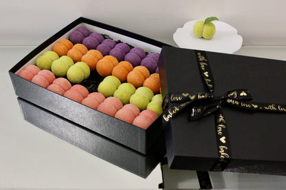 An open black gift box filled with rows of colorful macarons in pink, orange, yellow-green, and purple, next to a closed black box tied with a black ribbon printed with gold hearts and the words 'love you with all my heart'. A small white plate with two green fruits sits in the background on a light surface.
