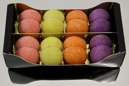 A black gift box containing 12 colorful sugar-coated macarons arranged in two rows of six. The macarons come in pink, yellow-green, orange, and purple flavors, set against a neutral light gray background.