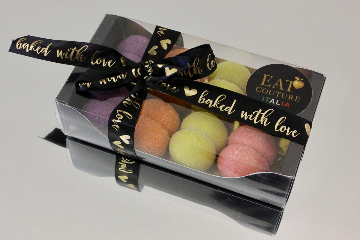 A clear plastic gift box containing several pastel-colored macarons, tied with a black ribbon printed with gold hearts and the words 'baked with love.' A round black sticker on the box reads 'EAT COUTURE ITALIA.