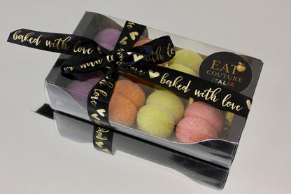 A clear plastic gift box containing several pastel-colored macarons, tied with a black ribbon printed with gold hearts and the words 'baked with love.' A round black sticker on the box reads 'EAT COUTURE ITALIA.