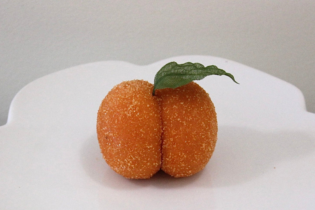 A white ceramic pedestal plate with a scalloped rim, holding a small orange decorative object with a green leaf on top, set against a neutral gray background.